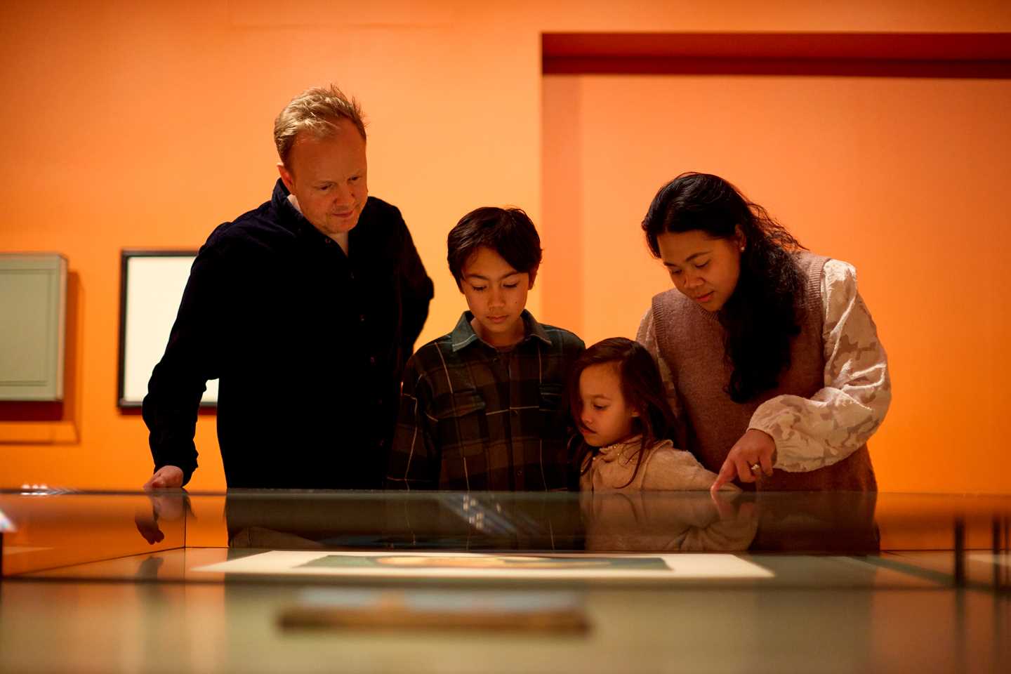 En familie på fire står ved et glassmontér og ser på et grafisk trykk av Edvard Munch. Fotografi