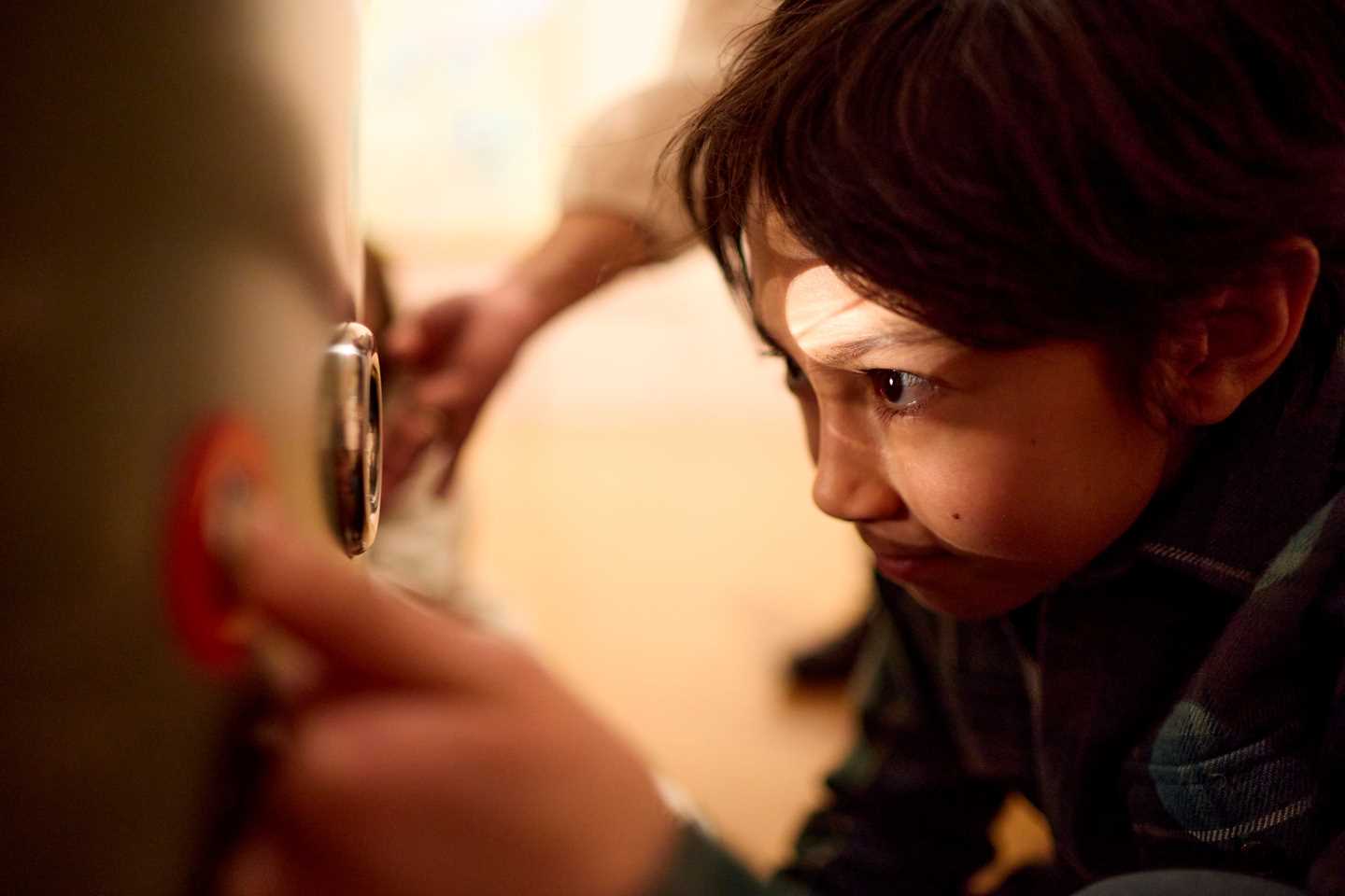 En ung gutt sitter på huk og stirrer inn i et kikkhull i en installasjon i utstillingen Monumental. Fotografi