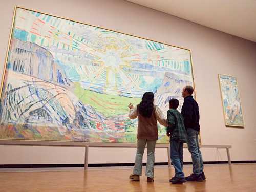 A mother, son and father stand looking at The Sun by Edvard Munch. They are facing away from the camera. Photograph.