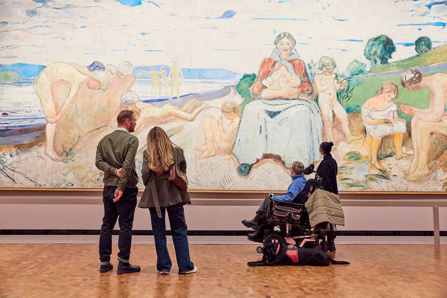 Fire personer står med ryggen mot kamera og betrakter maleriet Alma Mater av Edvard Munch. Bildet er 50 kvadratmeter stort. Fotografi