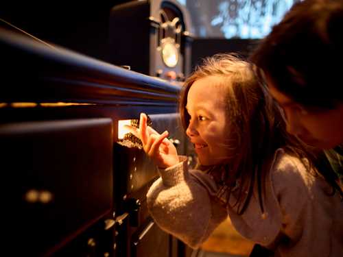 To barn huker seg ned og ser inn gjennom et musehull som det kommer lys ut av. Fotografi