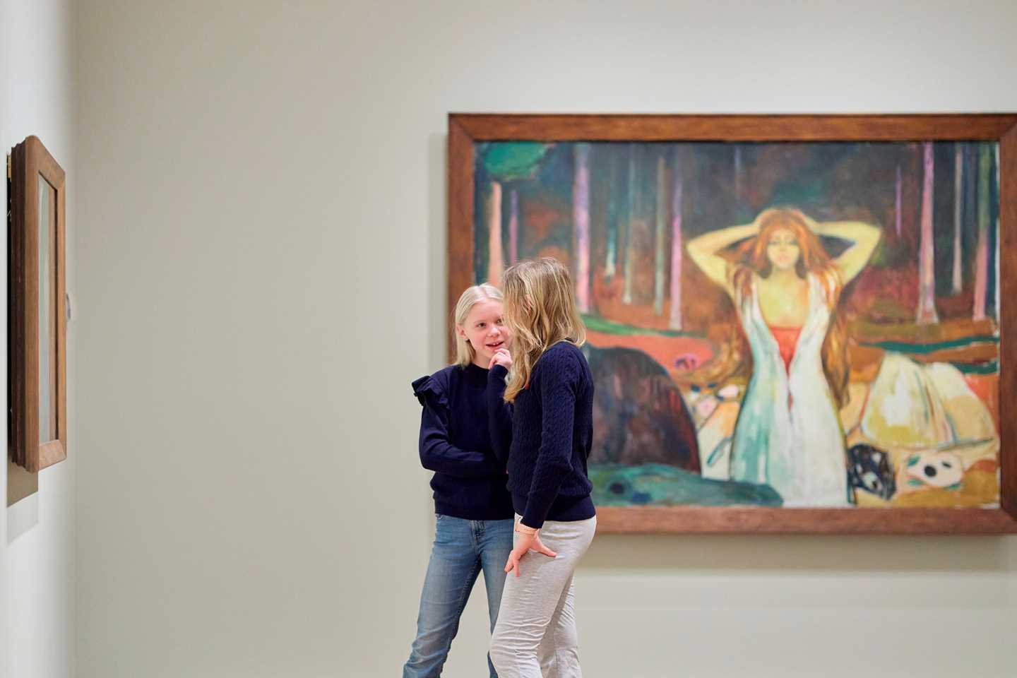 To unge jenter står og snakker sammen omgitt av kunstverk i utstillingen Uendelig. Fotografi