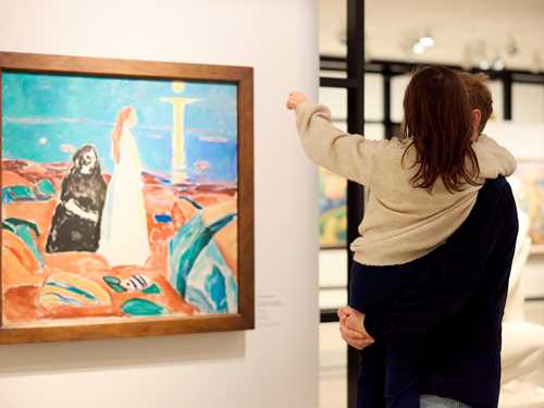 En far står med datteren sin på armen, begge har ryggen mot kamera. De ser på et maleri av Edvard Munch. Fotografi