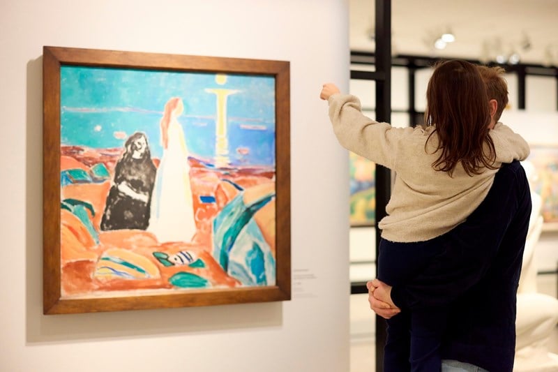 En far står med datteren sin på armen, begge har ryggen mot kamera. De ser på et maleri av Edvard Munch. Fotografi