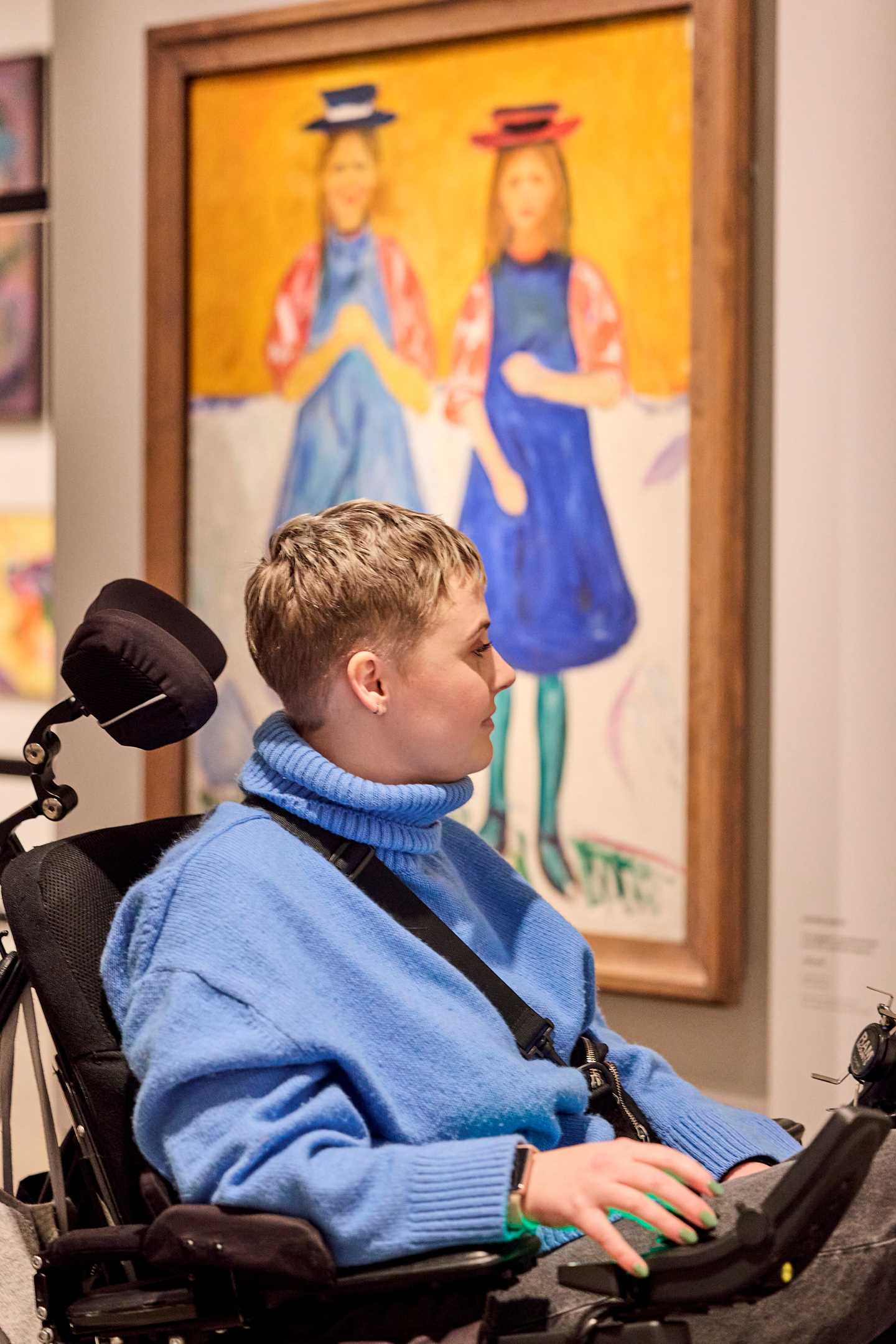 En kvinne i rullestol, sett fra siden, ser på et kunstverk. Bak henne ser vi verket To småpiker med blå forklær. Fotografi