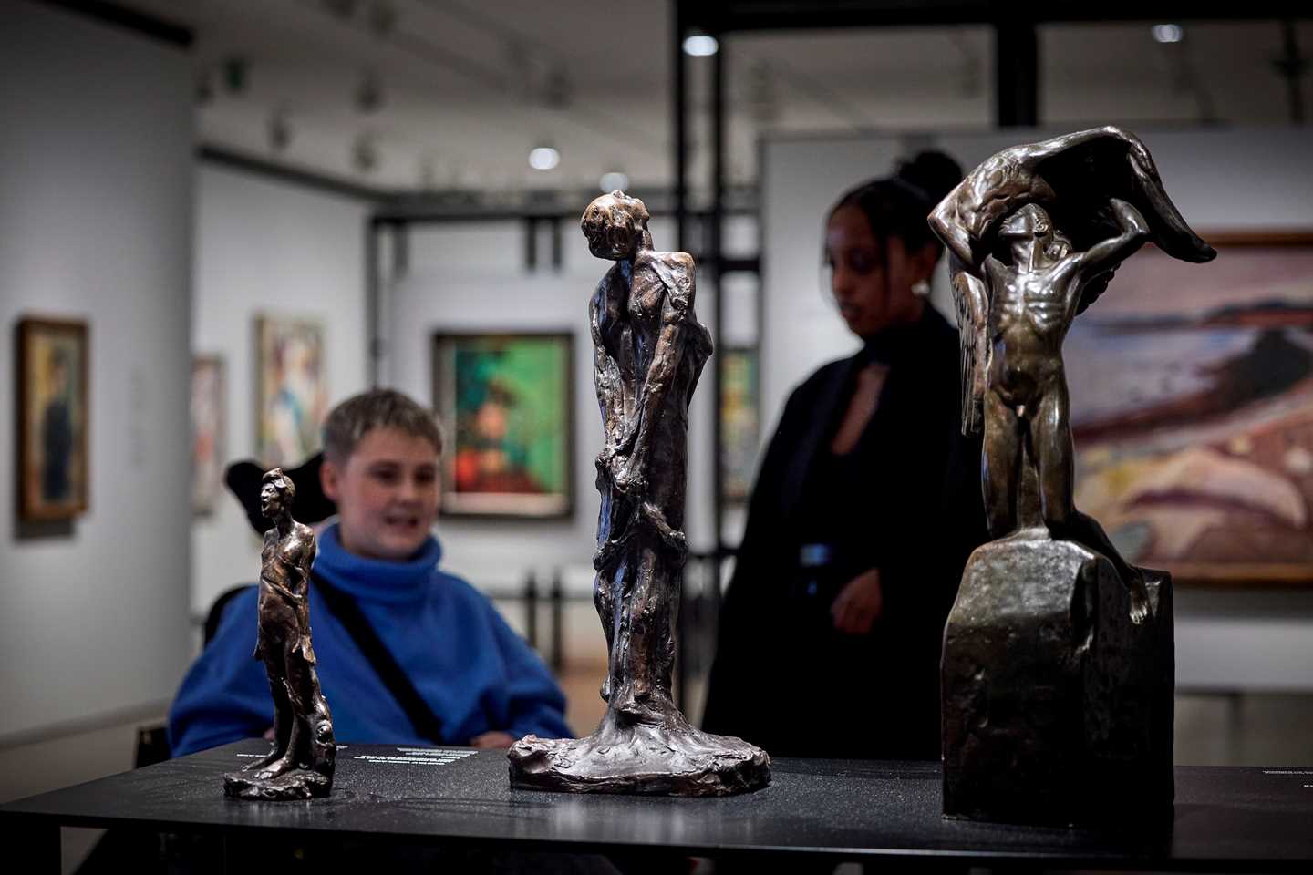 Tre bronseskulpturer av mennesker i ulike positurer. I bakgrunnen ser vi to personer i samtale og resten av utstillingsrommet. Fotografi