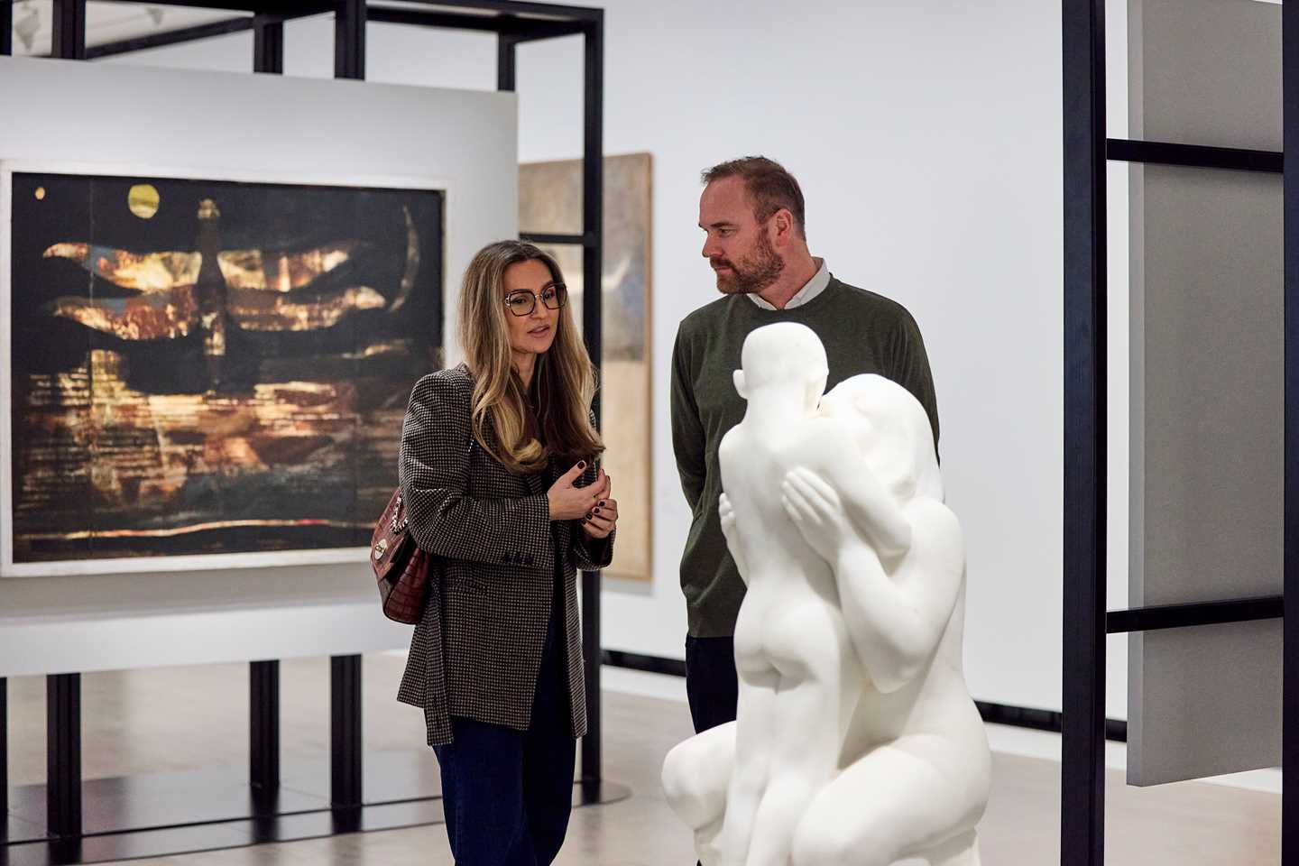En kvinne og en mann står i et utstillingsrom og har en samtale. Foran dem ser man en hvit skulptur av en kvinne og et barn. Bak dem ser man et kunstverk som henger på veggen. Fotografi