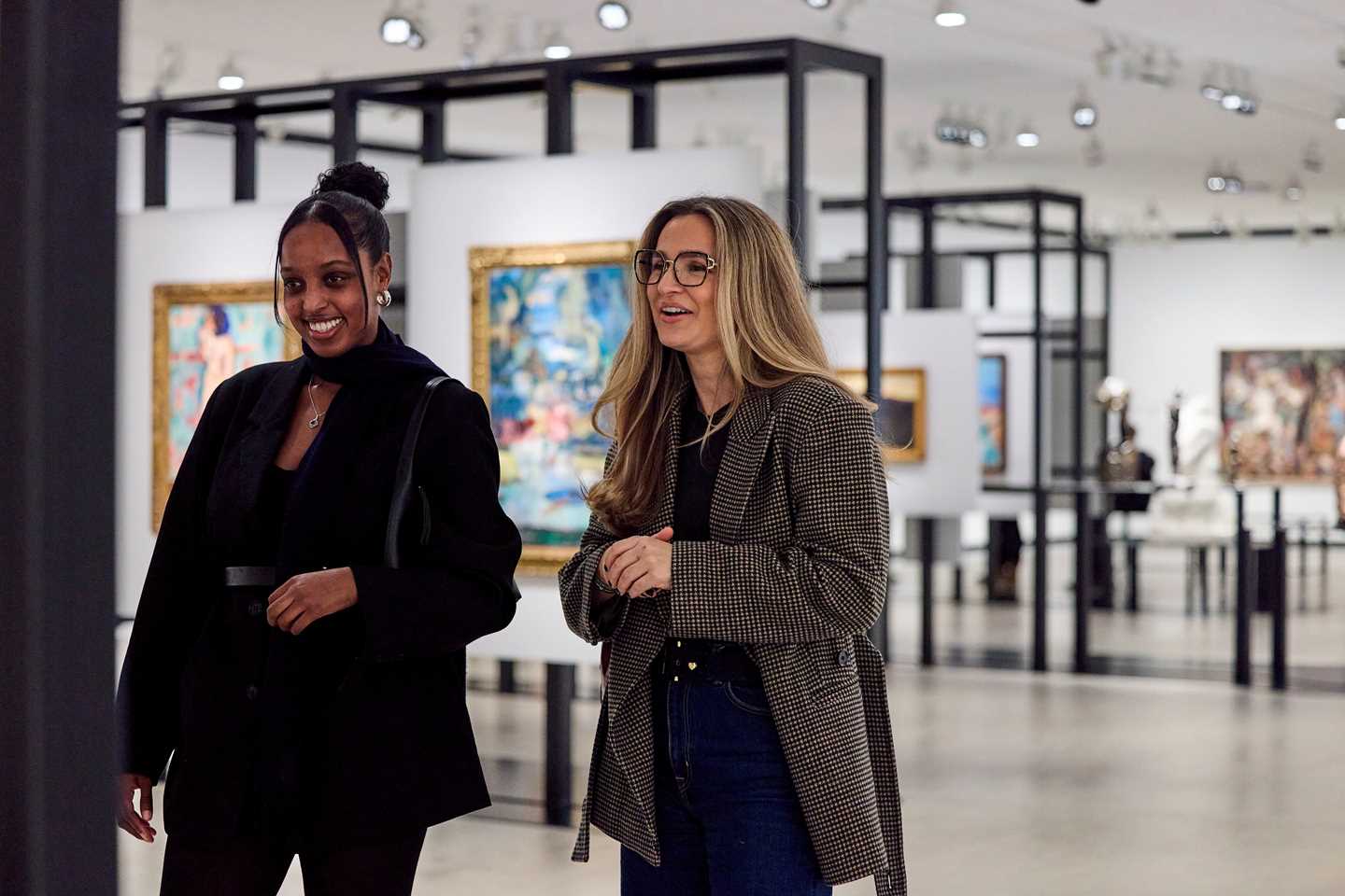 To kvinner står og ser på et kunstverk. Bak ser vi et utstillingsrom med svarte konstruksjoner som det henger kunstverk på. Fotografi