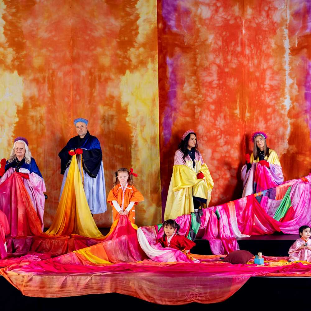 Fargerike stoffer henger ned i bakgrunnen og ligger drapert i forkant. Fem voksne og tre barn sitter og står inne stoffene. Foto