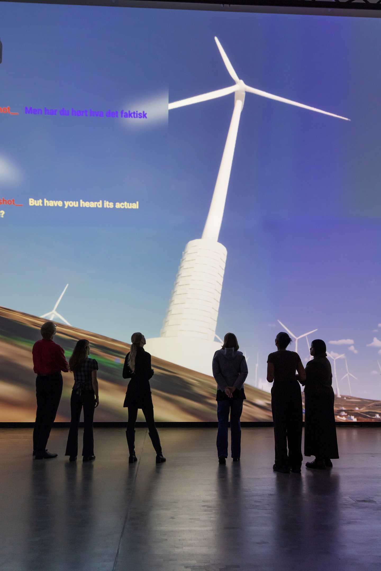 En gruppe mennesker står inne i et mørkt rom foran en vegg som er dekket av en projeksjon som viser en dataanimasjon av vindmøller på Golanhøydene, samt en boks med en chatdialog. Fotografi