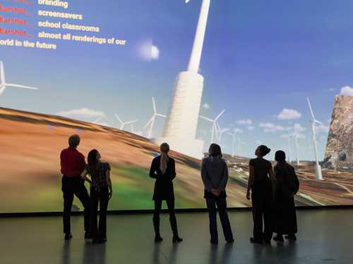 En gruppe mennesker står inne i et mørkt rom foran en vegg som er dekket av en projeksjon som viser en dataanimasjon av vindmøller på Golanhøydene. Fotografi