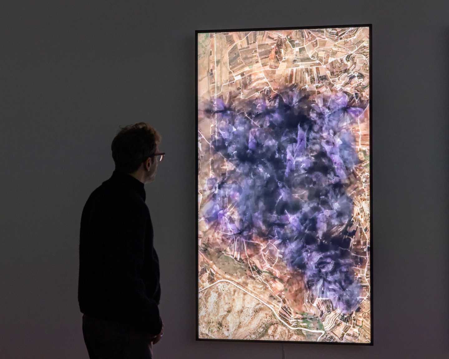 Kunstneren Lawrence Abu Hamdan står i et mørkt rom og ser på en lodrett skjerm som viser en visualisering av støyen 31 vindturbiner vil skape på Golanhøydene. På skjermen ser du at støyen sprer seg utover et stort område som lilla og svart gnistrende røyk. Fotografi