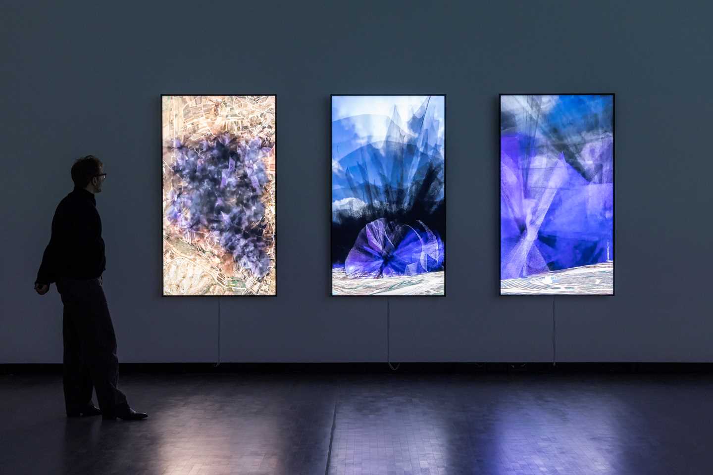 Kunstneren Lawrence Abu Hamdan står inne i et mørkt rom foran tre vertikale skjermer som henger på veggen og viser visualiseringer av støyen 30 vindturbiner vil skape på Golanhøydene. Støyen brer seg ut som sort og lilla røyk. Fotografi