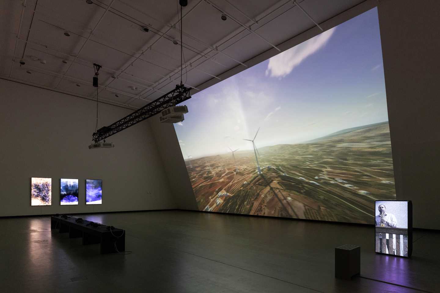 Et mørkt utstillingsrom som blir lyst opp av en projeksjon som dekker hele den ene veggen i rommet. Projeksjonen viser en dataanimasjon av vindturbiner på Golanhøydene. Midt i rommet står en svart benk og på veggen til venstre i bildet henger tre vertikale skjermer. Helt til høyre i bildet ser man en høyttaler med en projeksjon av saksofonisten Amr Mdah som spiller fra en balkong. Fotografi