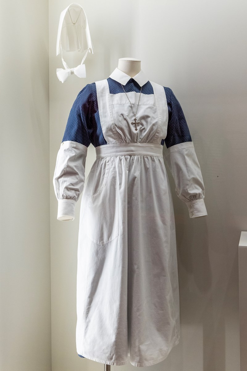 Diakonisseuniform på utstillingsdukke: hvit skjorte, blått skjørt med hvite prikker, hvitt forkle, løse hvite ermer, svarte strømpebukser og hvit sykepleierlue med hakestropp. Luens bruk markerte fullført utdanning. Fotografi