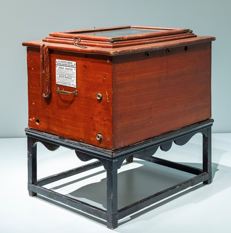 En kuvøse laget av mørkt beiset tre står på et stativ av svartbeiset tre som har utskjæringer i dekorative bølger. I lokket er det en glassplate som gjør at du kan se inn i kuvøsen.