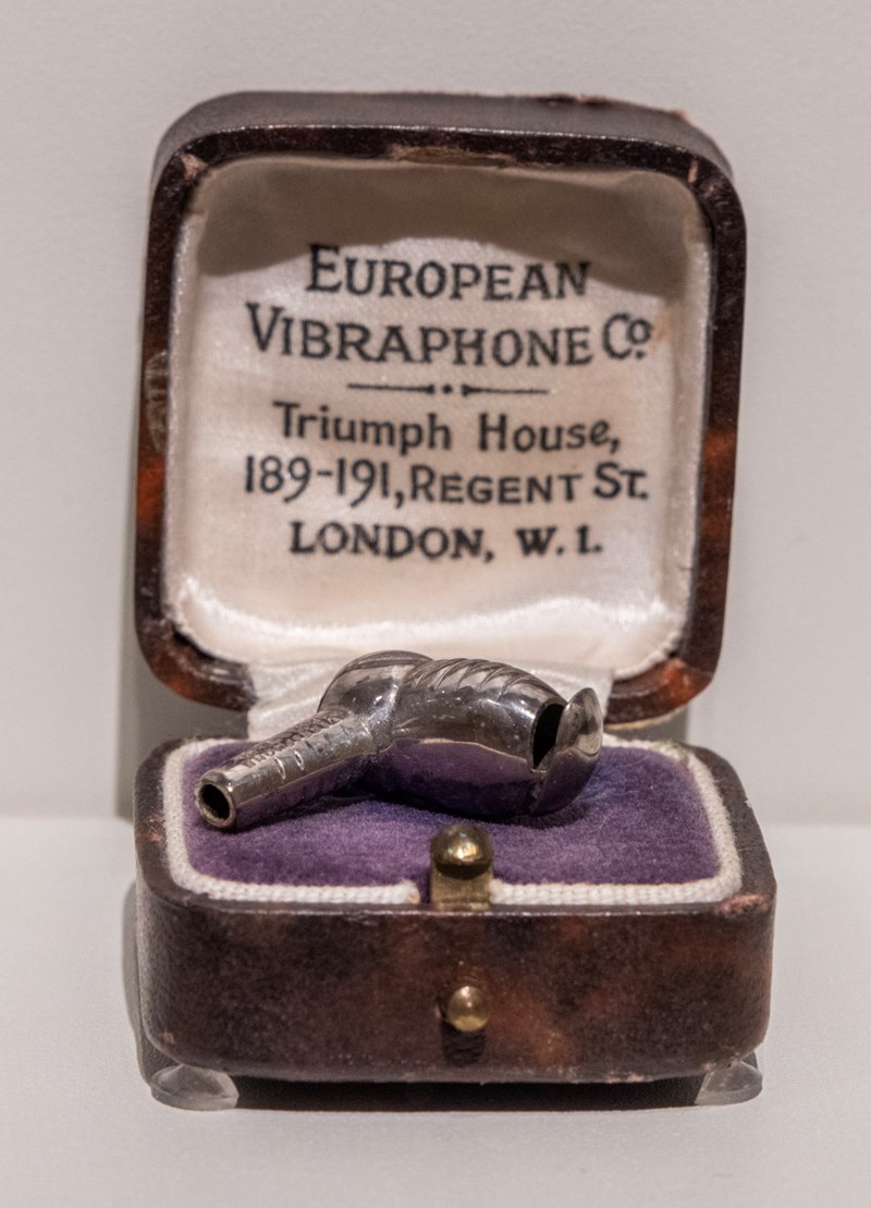 Et lite etui i lær, foret med et hvitt silkestoff. I bunnen av boksen ligger en indigofarget velurpute, hvor et høreapparat i sølv ligger.