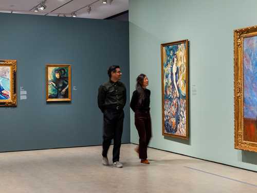 To mennesker går gjennom en utstilling og ser på malerier av Ludvig Karsten mens de smiler. Fotografi