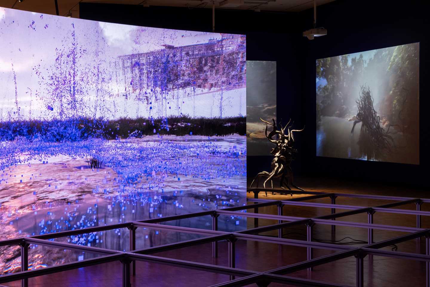 In the foreground, a curved screen displays a dystopian landscape. In the background, sculptures and accompanying video works by Sahej Rahal are visible. Photograph.