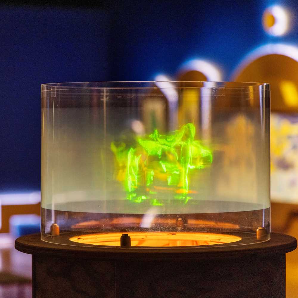A hologram showing dancing green figures is in focus. Behind it, a blue exhibition room and another art installation are visible. Photograph.