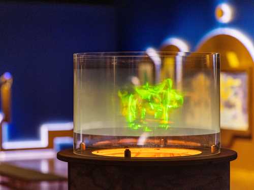 Et hologram som viser dansende, grønne mennesker er i fokus i bildet. Bak ser vi et blått utstillingsrom og en annen kunstinstallasjon. Fotografi