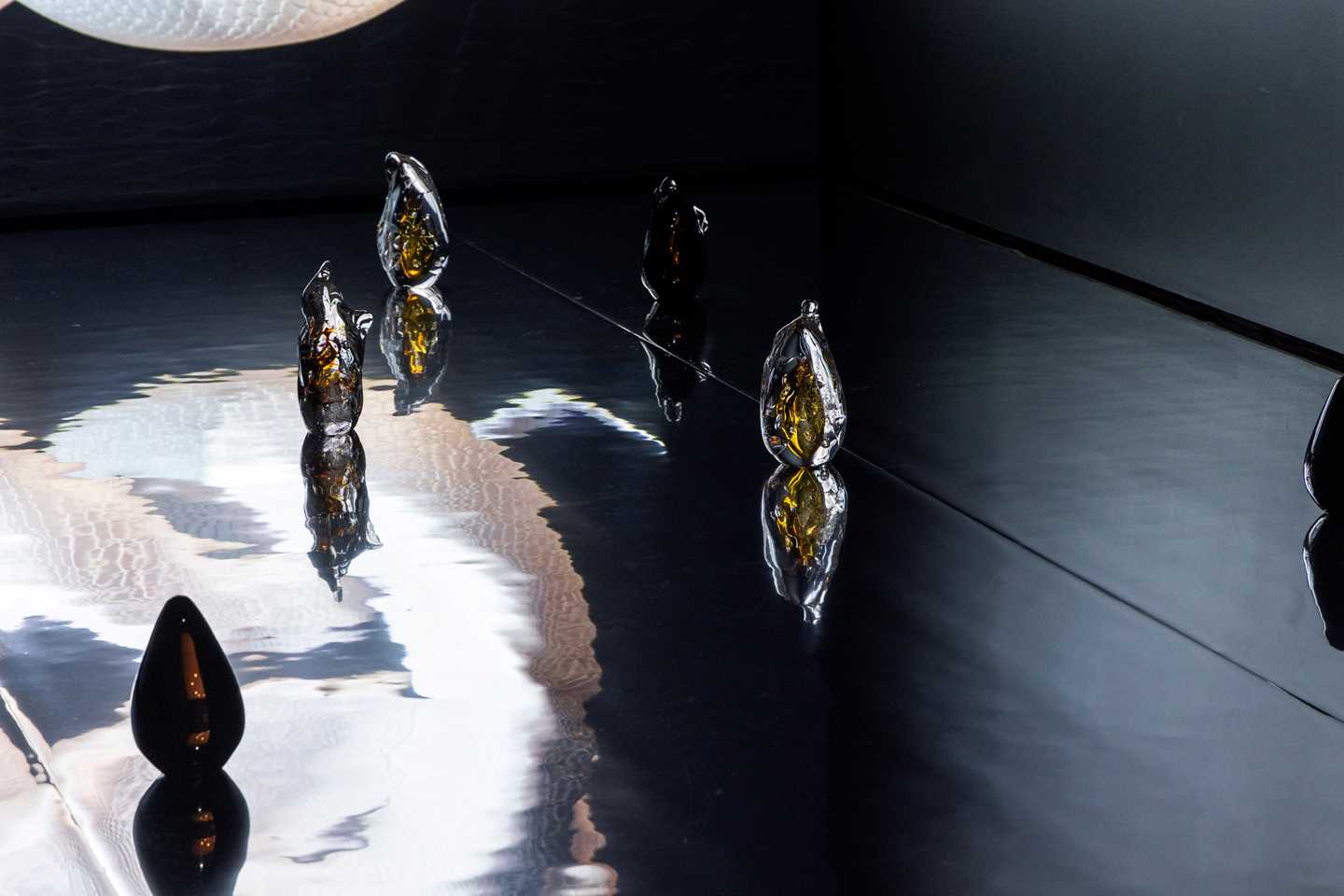 Close-up of glass sculptures standing on the floor, which is covered with a film that makes it resemble water, reflecting the sculptures. Photograph.