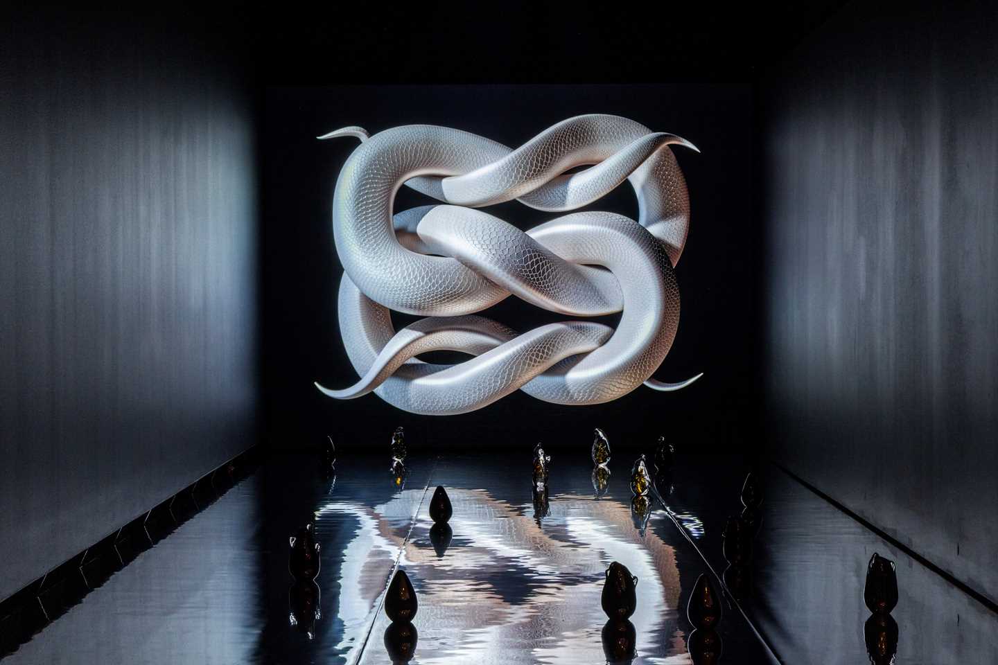 A dark, elongated room where a white, snake-like knot is displayed at the far end. The floor is covered with a glossy film reminiscent of a water surface, and on top of it stand glass sculptures. Photograph.
