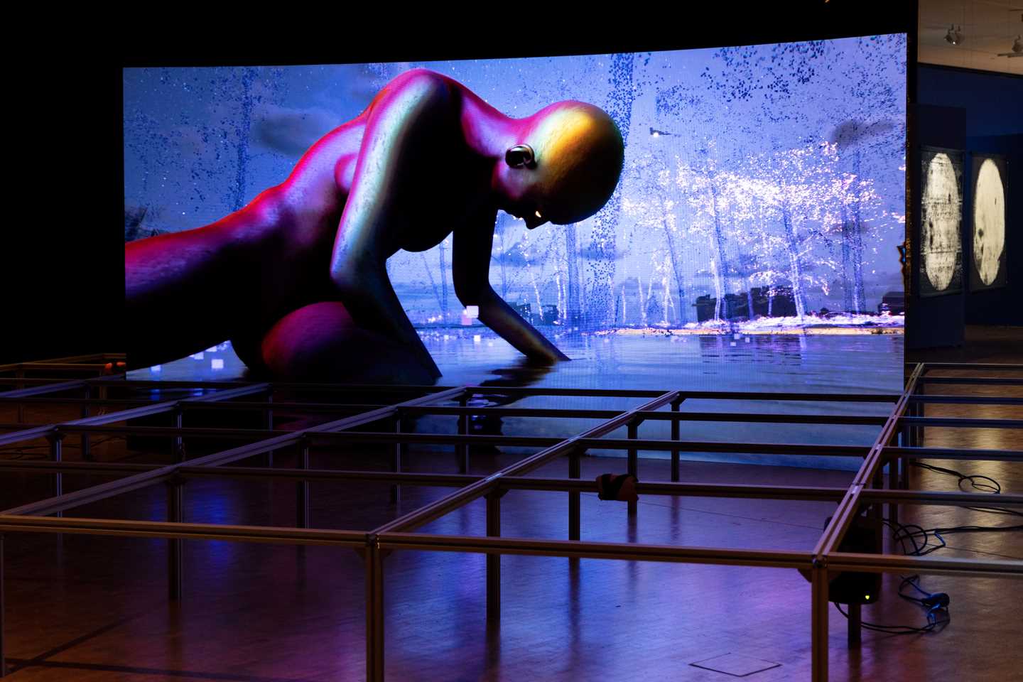 A large curved screen displays a computer-animated video, and on the floor around it stands a metal structure consisting of a grid fitted with speakers. The artwork is created by the artist collective Infopsin. Photograph.