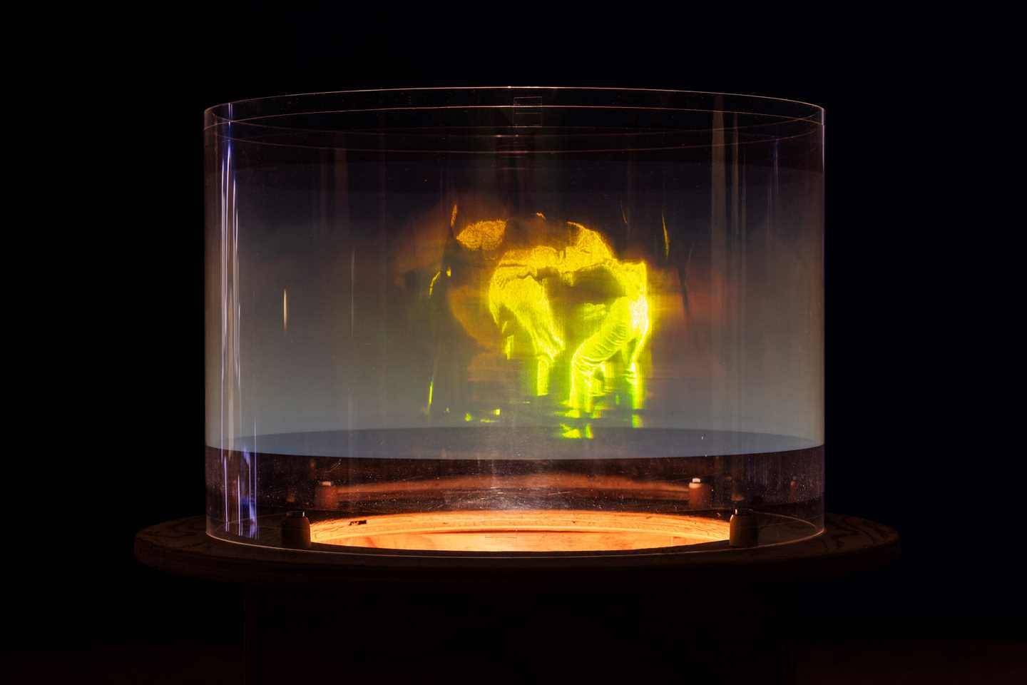 A hologram showing people dancing together in a circle. The figures are green, illuminating the otherwise dark image. Photograph.