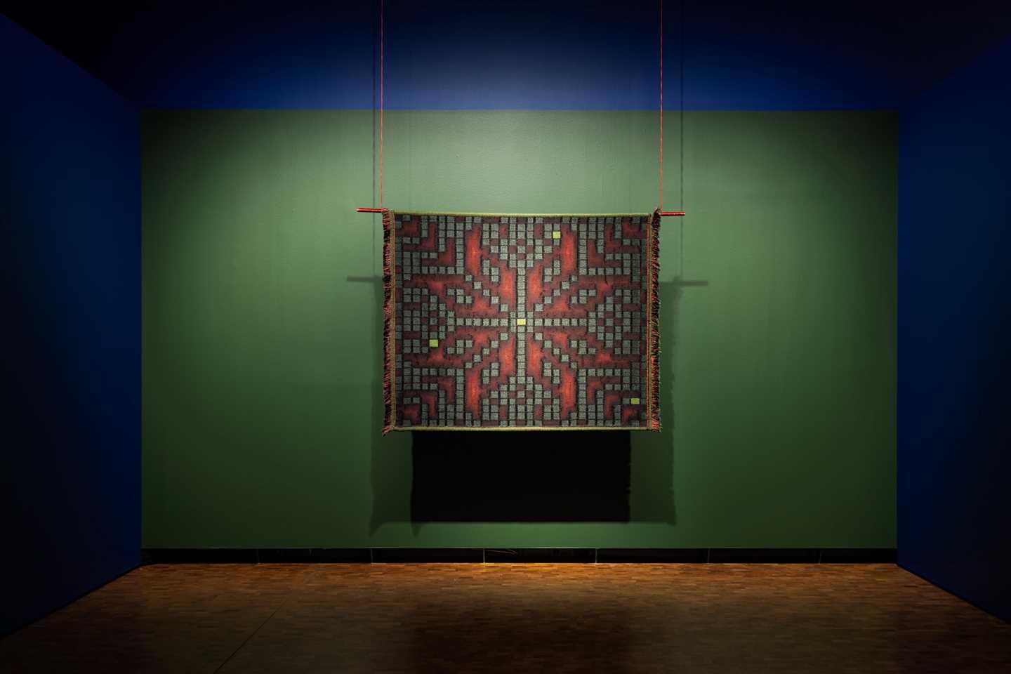 A square tapestry hangs against a green background. The pattern resembles the Selbu rose but is composed of small boxes that aesthetically resemble Tetris or similar video game blocks. Photograph.
