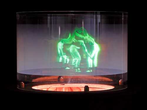 Et hologram viser mennesker som danser sammen i en ring. Menneskene er grønne og lyser opp bildet som ellers er helt mørkt. Fotografi