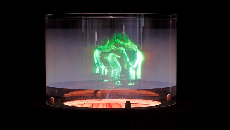 Et hologram viser mennesker som danser sammen i en ring. Menneskene er grønne og lyser opp bildet som ellers er helt mørkt. Fotografi