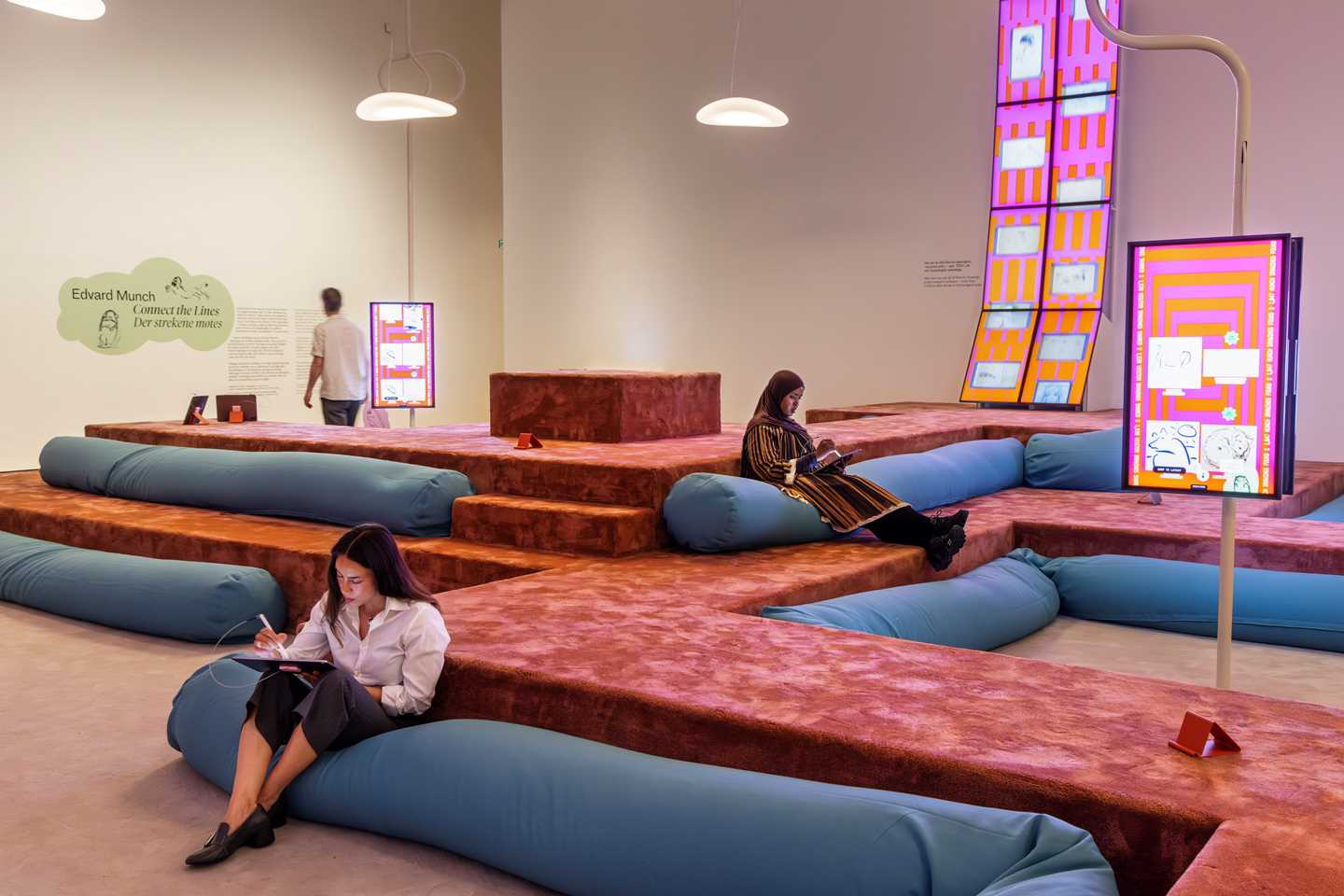 Et rom fylt av lange blå pølseputer som ligger rundt på et amfi laget av et mykt terracottafarget stoff. Rundt i rommet henger skjermer med sterke farger i rosa og oransje, som viser tegninger av Edvard Munch og tegninger skapt av publikum i rommet. To personer sitter på hver sin pølsepute og tegner på iPads, en tredje person står og leser en tekst på veggen. Fotografi