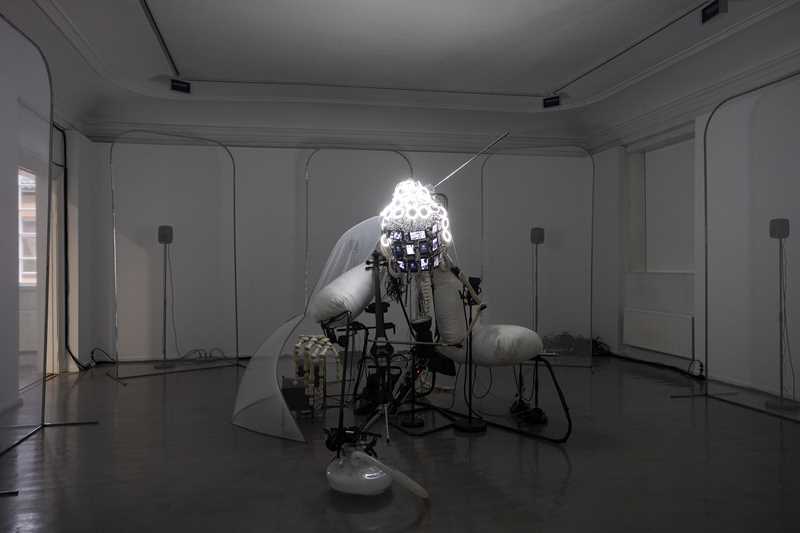 A dimly lit white room is primarily illuminated by an installation in the centre of the space, featuring an Buddha-like head covered in LEDs that emit a sharp white light. Photograph.