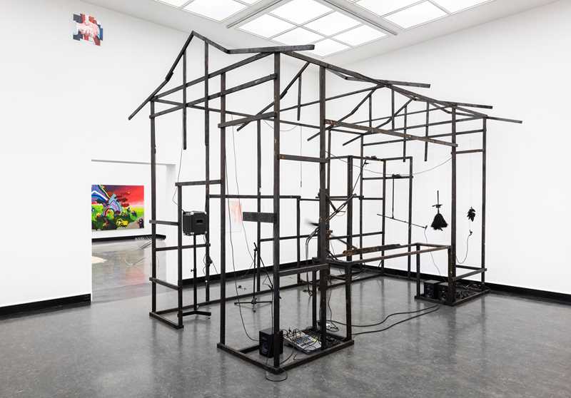 In a white gallery space, many black rods form the framework of a small, elongated house. Photograph.
