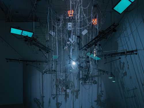 En installasjon henger ned fra taket i et mørkt rom lyst opp av blått lys. Installasjonen består blant annet av mange ledninger, strenger til musikkinstrumenter, lysskjermer og tastaturer. Fotografi