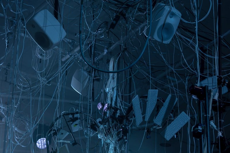 Høyttalere og tastaturer henger ned fra taket i et virrvarr av ledninger og strenger. Fotografi
