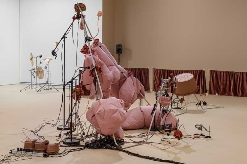 An installation in a room with sand-coloured and white walls. In the background, stands with cymbals are visible. In the foreground, stands fitted with pink fabric cushions can be seen. Wires run throughout, connecting different parts of the installation. Photograph.