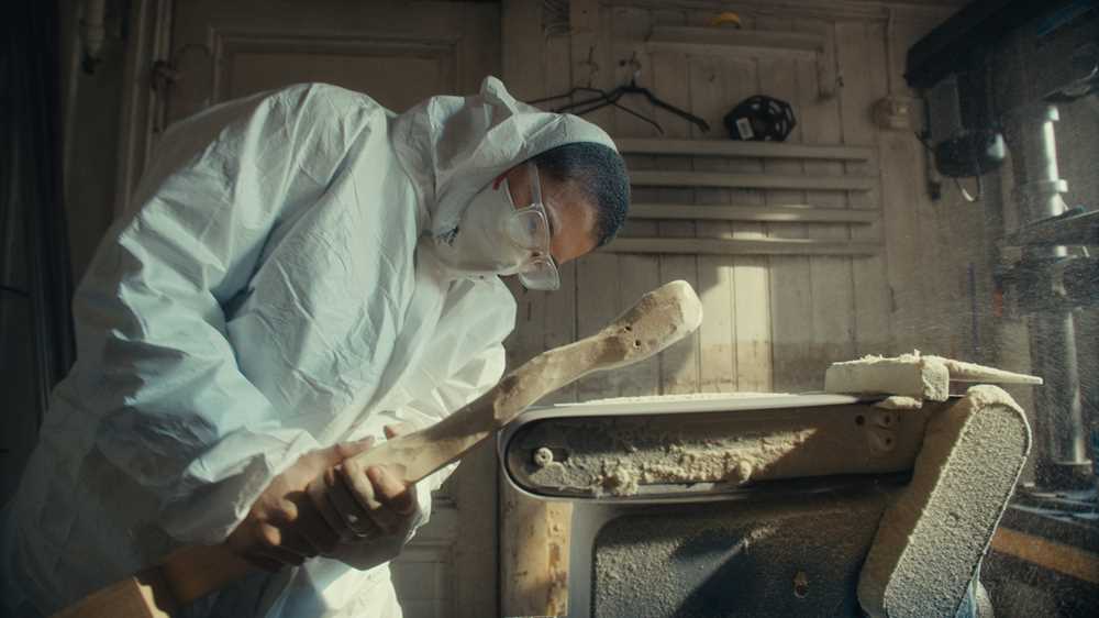 Kunstner Kim Hankyul i heldekkende hvit maledress, med munnbind og vernebriller står krumbøyd og sliper en trestokk på en automatisk slipemaskin. Fotografi