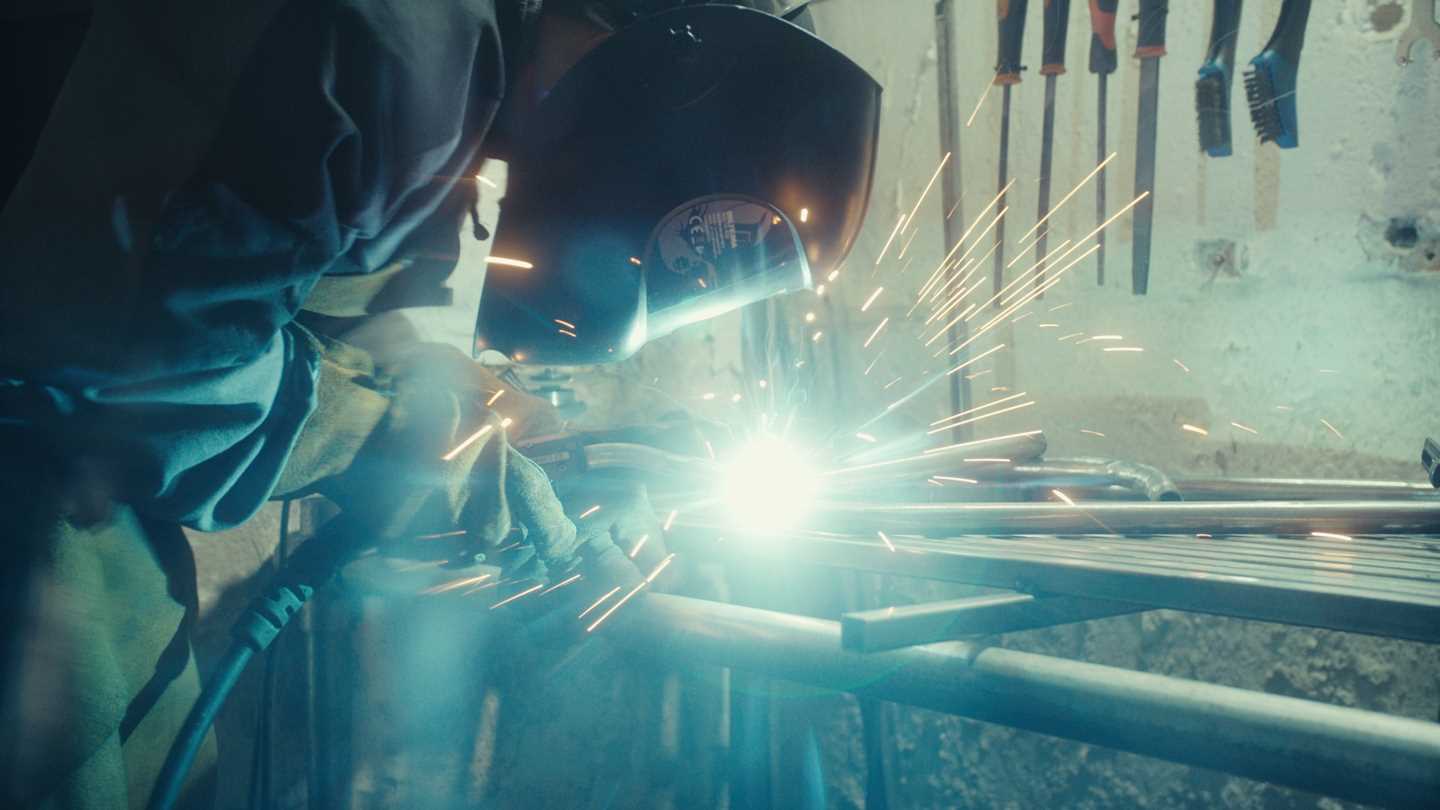 Artist Kim Hankyul bent over a workbench welding metal, wearing a protective visor with bright light and sparks around his face. Photograph.