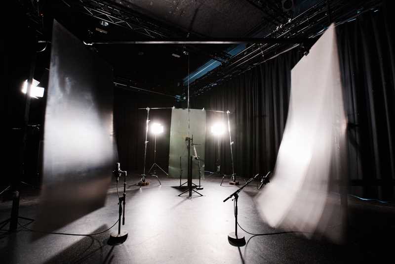 A dark room lit by two spotlights placed on the floor and pointing directly towards the camera. On either side of the room, two thin panels hang from the ceiling. In front of each panel stands a microphone, and one of the panels is vibrating back and forth, made visible by the long exposure of the photograph, which captures the movement. Photograph.