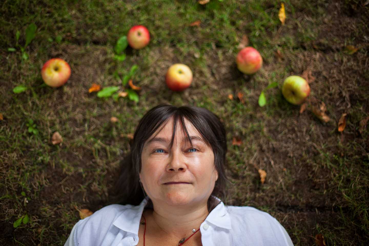 A photograph of the author Ali Smith lying on her back on the grass. The image is taken directly from above, and Ali looks straight up at the camera. On the ground above her head are five apples, as if they have fallen from an apple tree. Photograph.