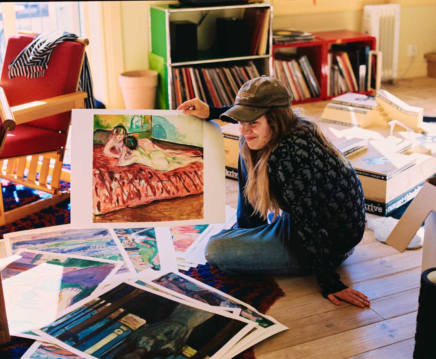 Artisten Marie Ulven sitter på gulvet i en leilighet med masse plakater som viser verk av Edvard Munch rundt seg. Marie har på seg caps og holder opp et verk av to nakne kvinner på en seng. Hun ser ut som hun skal til å begynne å le. Fotografi