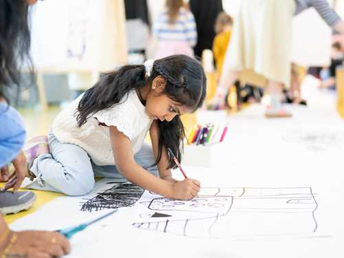 A girl sits on the floor drawing on a large sheet of paper taped to the floor. She is deeply absorbed in her work. Photograph.
