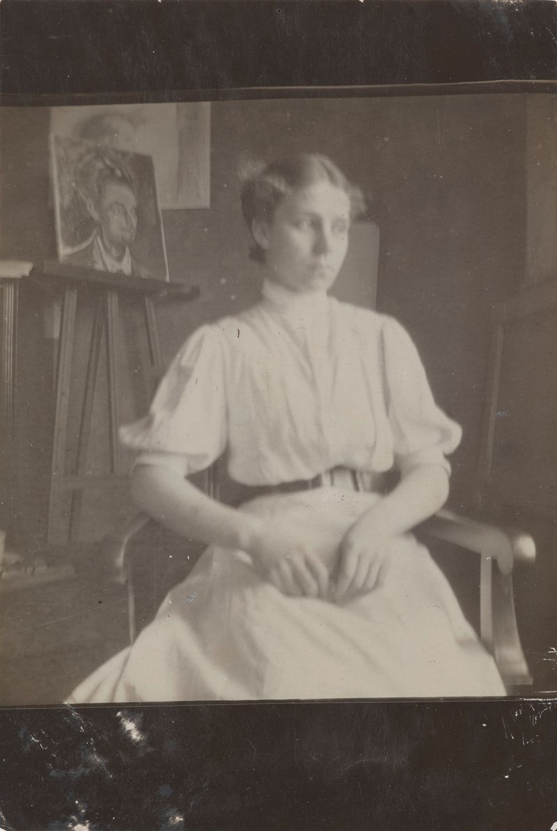 En kvinne i hvit kjole sitter med hendene i fanget og blikket vendt bort. I bakgrunnen vises Munchs atelier med et maleri på veggen og ett på staffeli. Fotografi