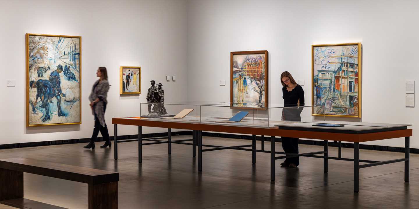 Two women in an exhibition gallery. One is looking at a painting, while the other studies something displayed in a glass case. Photograph.