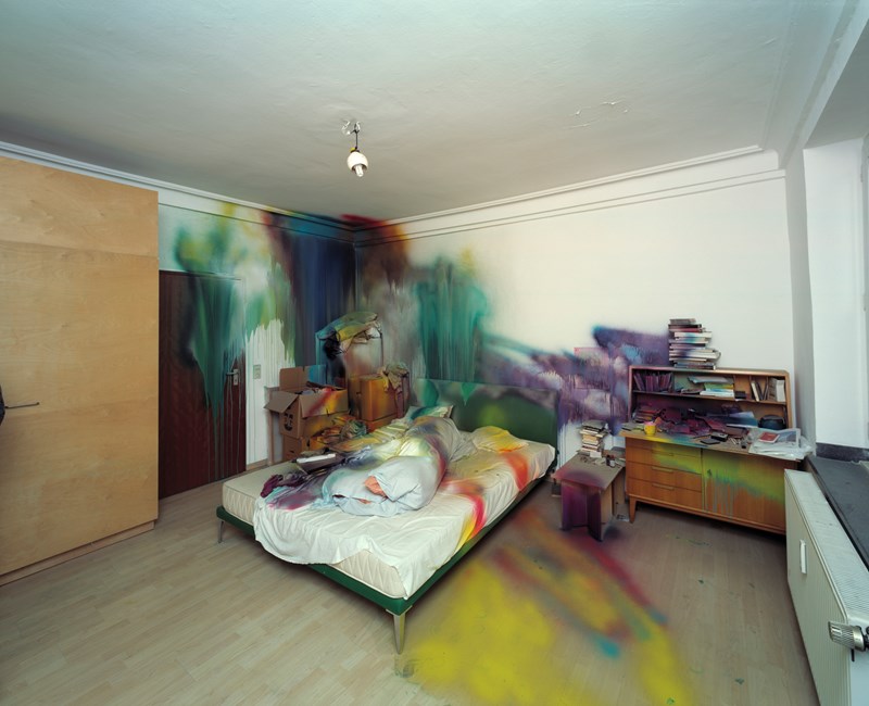 Et rom med en seng midt i og diverse møbler på hver side av sengen. Sengen, deler av rommet og møblene er spraymalet i ulike farger. Fotografi