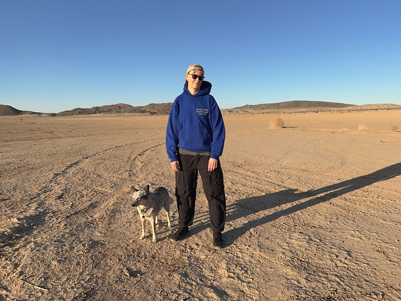Vi ser en person i helfigur med blå hettegenser, svarte bukser og solbriller stående i et ørkenlandskap med en hund ved siden av seg. I bakgrunnen er det fjell og himmelen er knallblå. Fotografi