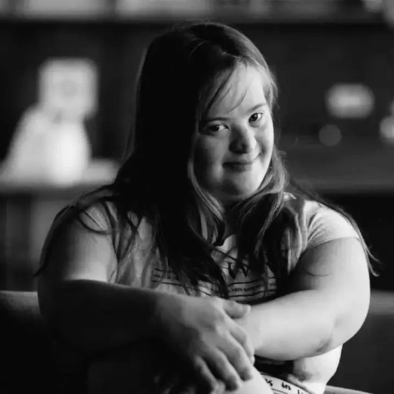 A black-and-white photo of a smiling woman with loose hair, wearing a short-sleeved top, seated cross-legged in an armchair with hands on her knees. Shelves with framed photos and objects are behind her.