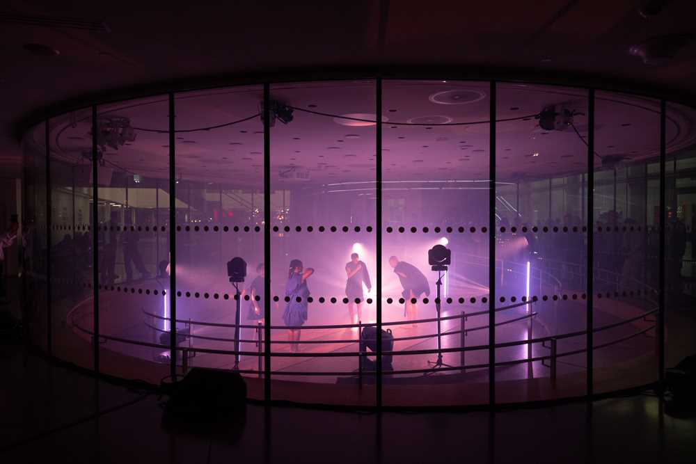 Fire mennesker danser inne i et glassrom lyst opp av lyskastere med lilla lys. Fotografi